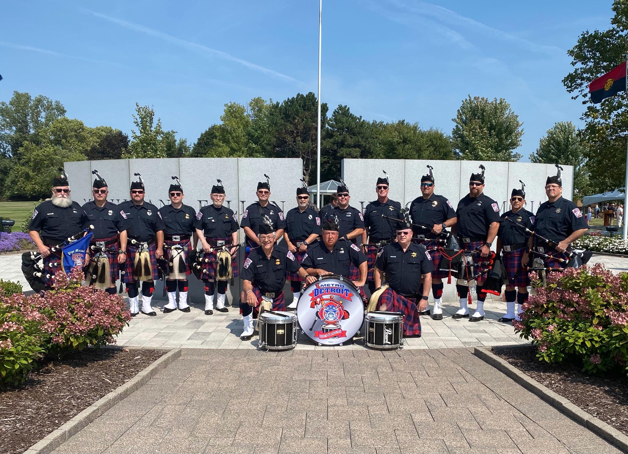 Members of the Metro Detroit Police and Fire Pipes and Drums in full dress, standing before a fallen heroes memorial wall